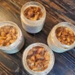 Four jars of apple pie overnight oats topped with cinnamon-spiced apples on a wooden table
