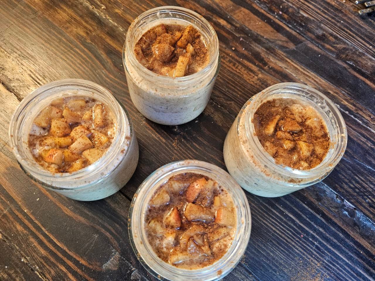 Four jars of apple pie overnight oats topped with cinnamon-spiced apples on a wooden table